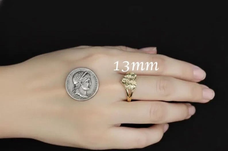 Puede incluir: Un anillo dorado con un grupo de piedras se muestra junto a una moneda. El anillo mide aproximadamente 1,3 cm. El anillo est&aacute; en un dedo. El fondo es negro.