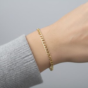 May include: A gold-colored curb chain bracelet is worn on a wrist. The bracelet is made of small, interlocking links. The person is wearing a gray sweater. The background is a solid, light gray.
