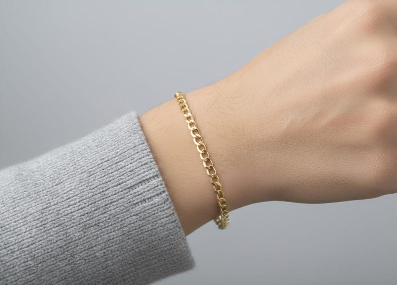 May include: A gold-colored curb chain bracelet is worn on a wrist. The bracelet is made of small, interlocking links. The person is wearing a gray sweater. The background is a solid, light gray.