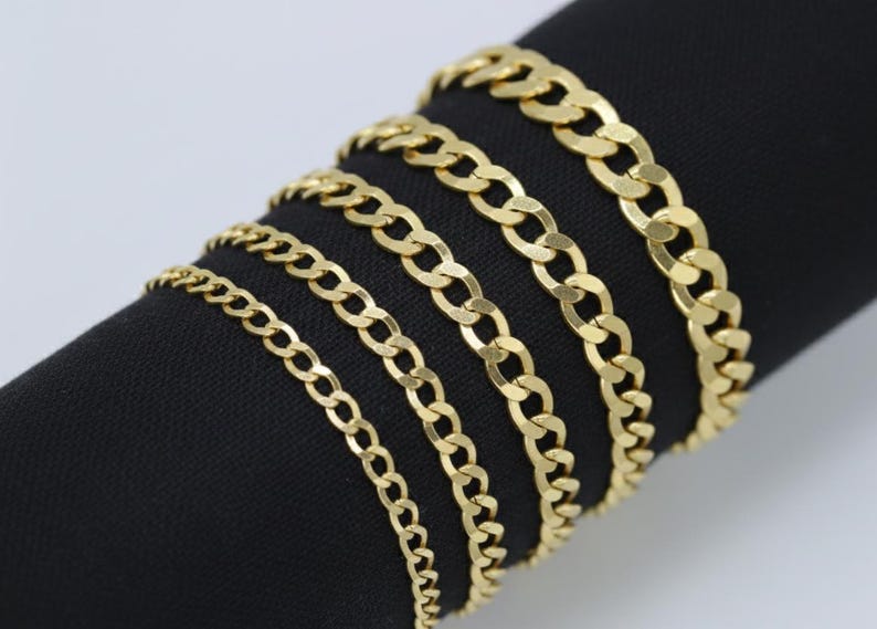 May include: A collection of gold-colored curb chains of varying thicknesses, displayed on a black surface. The chains are arranged side by side, showcasing the different sizes and styles of the jewelry. The chains are made of a shiny, reflective metal.