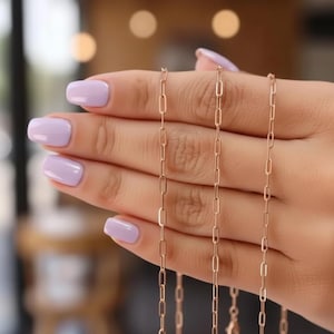 Könnte beinhalten: Drei zarte goldfarbene Kettenhalsketten mit einem Büroklammer-Glieder-Design werden gezeigt. Die Ketten werden vor einem verschwommenen Hintergrund gehalten. Die Glieder sind länglich und rechteckig. Die Ketten haben eine warme, goldene Farbe.