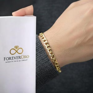 May include: A gold curb link bracelet is worn on a wrist. A white box with the logo "FOREVER ORO" and the text "AUTHENTIC 10K & 14K JEWELRY" is held in the other hand. The bracelet is a classic jewelry design.