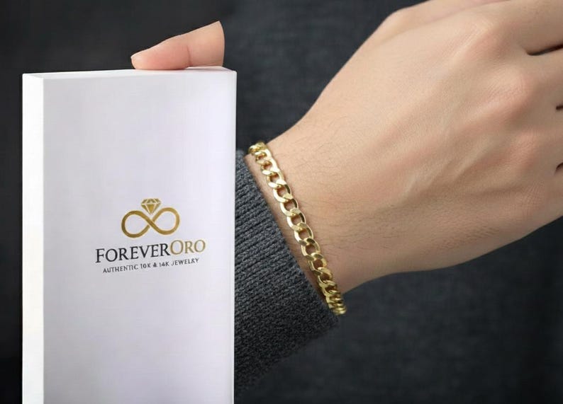 May include: A gold curb link bracelet is worn on a wrist. A white box with the logo "FOREVER ORO" and the text "AUTHENTIC 10K & 14K JEWELRY" is held in the other hand. The bracelet is a classic jewelry design.