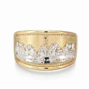 Pode incluir: Um anel dourado com um relevo detalhado da Última Ceia. O anel tem uma banda larga com uma superfície texturizada e uma cruz no topo. As figuras são em metal prateado, criando um efeito de duas cores.
