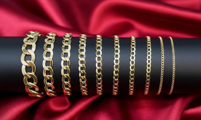 May include: A collection of gold-colored chain bracelets displayed on a black cylindrical stand against a red satin backdrop. The bracelets vary in chain link size, showcasing different styles and widths. The chains are arranged in a row, highlighting the variety.