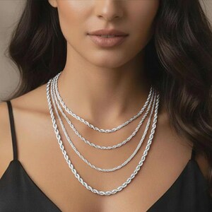 Puede incluir: Un conjunto de cuatro collares de cadena de cuerda plateados de diferentes longitudes. Los collares se muestran sobre un fondo oscuro, resaltando su intrincado dise&ntilde;o trenzado. Las cadenas son un elemento b&aacute;sico de la joyer&iacute;a cl&aacute;sica.
