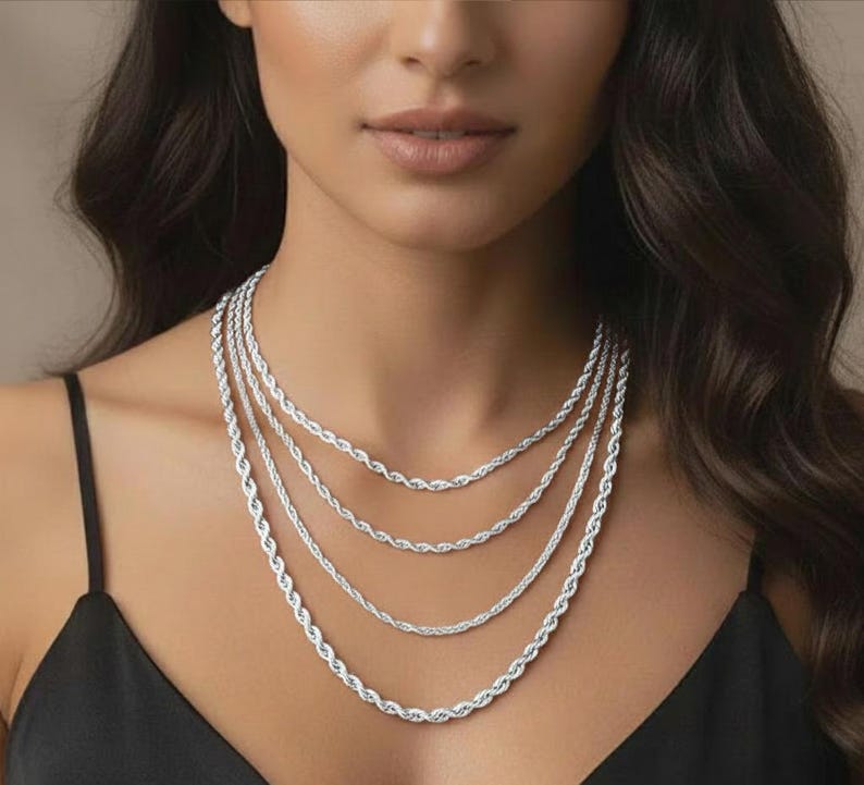 Puede incluir: Un conjunto de cuatro collares de cadena de cuerda plateados de diferentes longitudes. Los collares se muestran sobre un fondo oscuro, resaltando su intrincado dise&ntilde;o trenzado. Las cadenas son un elemento b&aacute;sico de la joyer&iacute;a cl&aacute;sica.