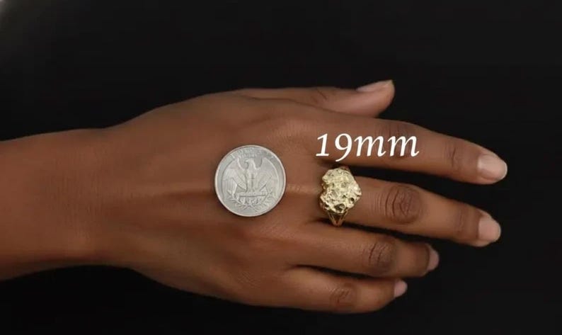 Puede incluir: Un anillo dorado con un dise&ntilde;o org&aacute;nico texturizado, junto a una moneda para comparar el tama&ntilde;o. El anillo tiene aproximadamente 19 mm de di&aacute;metro. Fondo oscuro.