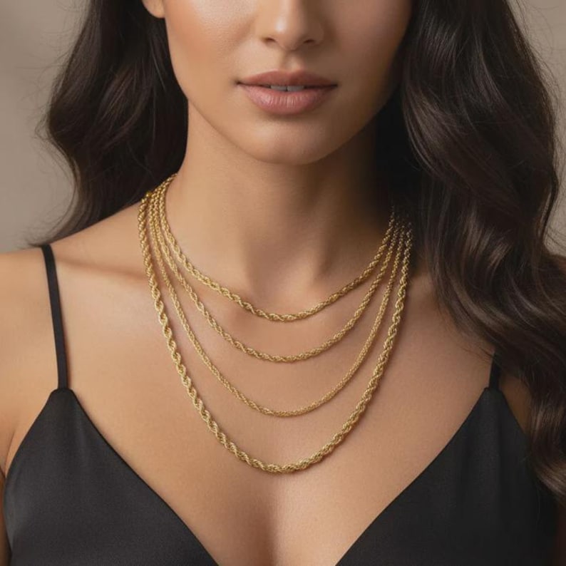 May include: A close-up of a person wearing four gold rope chain necklaces. The necklaces vary in thickness and length, creating a layered effect. The chains are a warm, shiny gold color, and the person is wearing a black top.