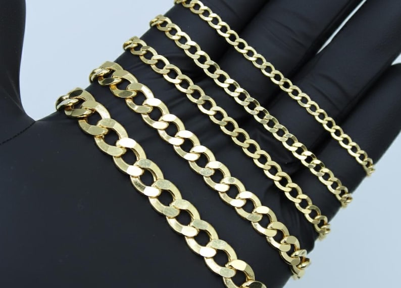 May include: A collection of gold-colored curb chain necklaces of varying widths. The chains are displayed on a black surface, showcasing their different link sizes and overall shine. The chains are a classic jewelry style.