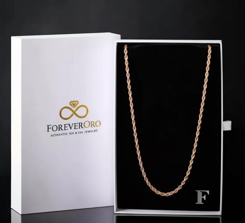 K&ouml;nnte beinhalten: Eine goldene Seilkette, pr&auml;sentiert in einer offenen wei&szlig;en Schachtel mit schwarzem Innenraum. Auf der Schachtel befinden sich das Logo und der Text "ForeverOro". Die Halskette ist mittig in der Schachtel platziert.