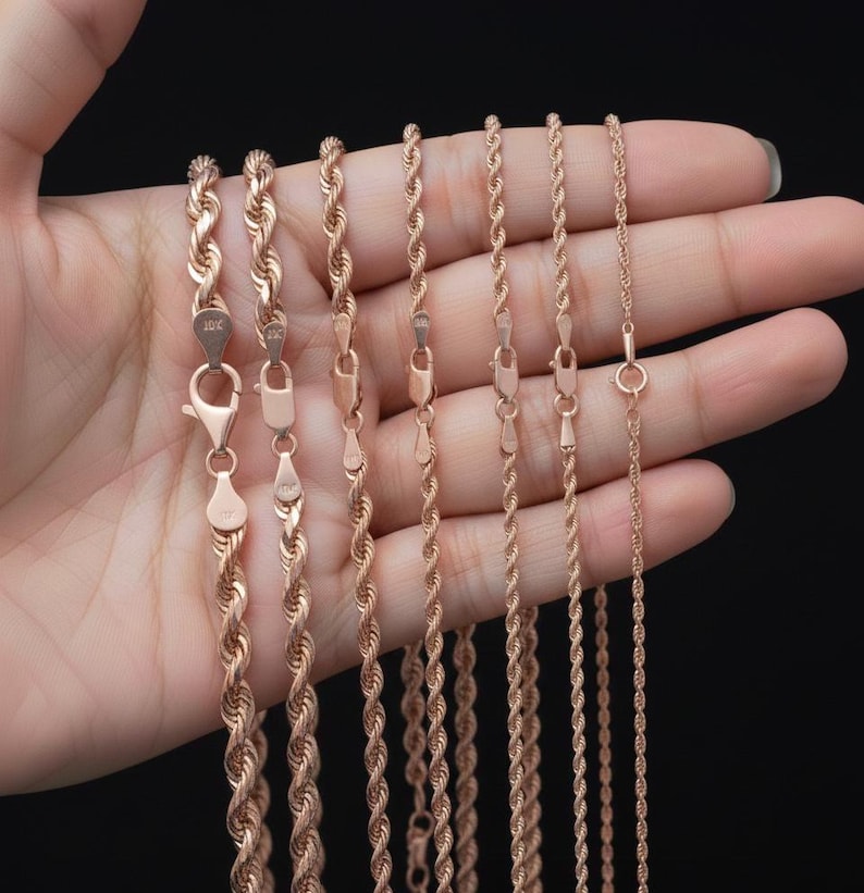 K&ouml;nnte beinhalten: Gezeigt werden mehrere Halsketten mit gedrehten Seilketten aus Ros&eacute;gold in verschiedenen St&auml;rken. Die Halsketten werden in einer Hand vor schwarzem Hintergrund gehalten. Jede Kette hat einen Karabinerverschluss. Die Ketten bestehen aus gl&auml;nzendem Metall.