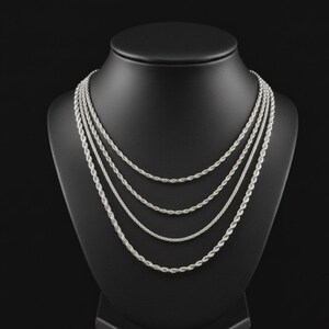 Puede incluir: Conjunto de collares de cadena de cuerda plateada exhibido en un maniqu&iacute; negro. El collar presenta m&uacute;ltiples hilos de diferentes longitudes, creando un efecto en capas. Las cadenas tienen un dise&ntilde;o de cuerda trenzada, que refleja la luz. El fondo es negro.
