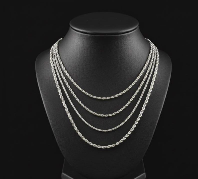 Puede incluir: Conjunto de collares de cadena de cuerda plateada exhibido en un maniqu&iacute; negro. El collar presenta m&uacute;ltiples hilos de diferentes longitudes, creando un efecto en capas. Las cadenas tienen un dise&ntilde;o de cuerda trenzada, que refleja la luz. El fondo es negro.