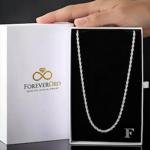 Puede incluir: Un collar de cadena de cuerda plateada exhibido en una caja blanca con el logotipo de ForeverOro. La caja est&aacute; abierta, revelando el collar sobre un interior negro. El collar tiene un dise&ntilde;o delicado y trenzado.