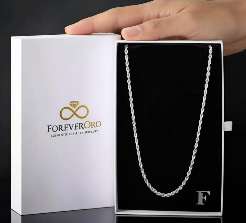 Puede incluir: Un collar de cadena de cuerda plateada exhibido en una caja blanca con el logotipo de ForeverOro. La caja est&aacute; abierta, revelando el collar sobre un interior negro. El collar tiene un dise&ntilde;o delicado y trenzado.
