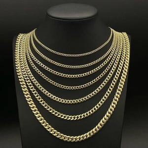 May include: A display of gold-colored curb chain necklaces of varying thicknesses. The necklaces are arranged on a black mannequin bust, showcasing the different sizes and styles. The chains have a shiny, polished finish.
