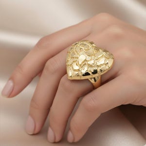 Könnte beinhalten: Ein auffälliger, goldfarbener Ring mit herzförmigem Design. Das Herz ist mit unregelmäßigen, sich überlappenden Formen strukturiert. Der Ring wird an einer Hand gezeigt, um seine Größe und die komplizierten Details des Designs hervorzuheben.