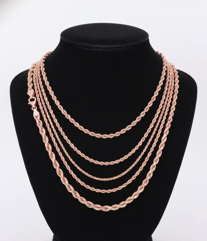 K&ouml;nnte beinhalten: Eine Sammlung von Halsketten mit Seilkette in Ros&eacute;gold, pr&auml;sentiert auf einer schwarzen Mannequin-B&uuml;ste. Die Halsketten variieren in der L&auml;nge und zeigen einen mehrschichtigen Look. Die Ketten haben ein gedrehtes Seildesign und einen Karabinerverschluss.
