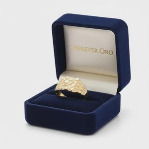 Puede incluir: Un anillo dorado con superficie texturizada, presentado en un estuche de terciopelo azul oscuro. El interior del estuche es blanco, con las palabras "FOREVER ORO" impresas en dorado. El anillo, con dise&ntilde;o irregular, es el foco principal.