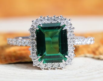 May include: A ring featuring a large, emerald-cut green centre stone surrounded by a rectangular halo of small, round, clear stones. The band is thin and also set with small, round, clear stones along the visible portion of the shank.