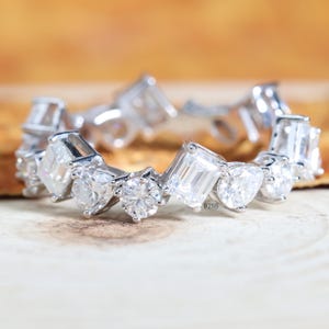 Può includere: Un anello eternity color argento con un motivo ripetitivo di pietre chiare che alternano il taglio smeraldo e il taglio rotondo su una fascia metallica. L'anello mostra il piccolo testo 218 vicino al centro, con gemme sfaccettate.