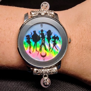 May include: Silver-toned wristwatch featuring a holographic dial with silhouettes of anime characters against a rainbow gradient. The watch has ornate detailing on the band and a decorative charm. A unique accessory.