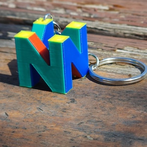 May include: A green, blue, yellow, and red 3D printed keychain shaped like the Nintendo logo. The keychain is attached to a silver metal ring.