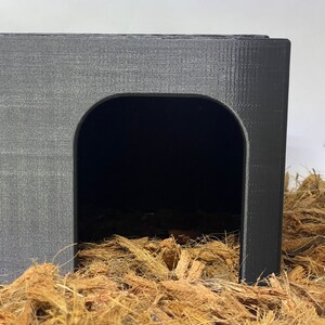 May include: A black plastic hide for small animals, featuring a rounded entrance and a textured surface. The hide is sitting on a bed of brown coconut fiber.