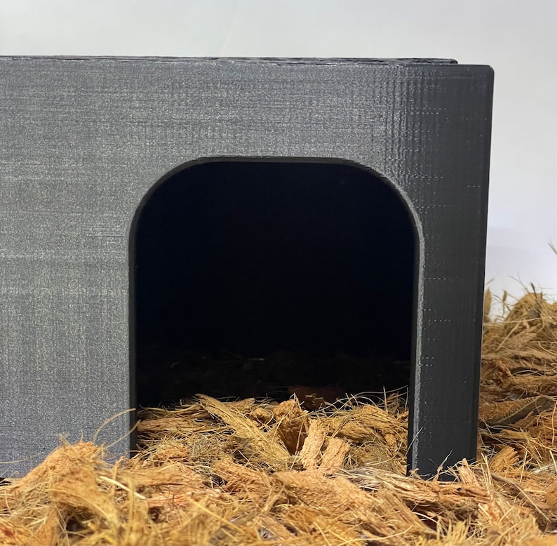May include: A black plastic hide for small animals, featuring a rounded entrance and a textured surface. The hide is sitting on a bed of brown coconut fiber.