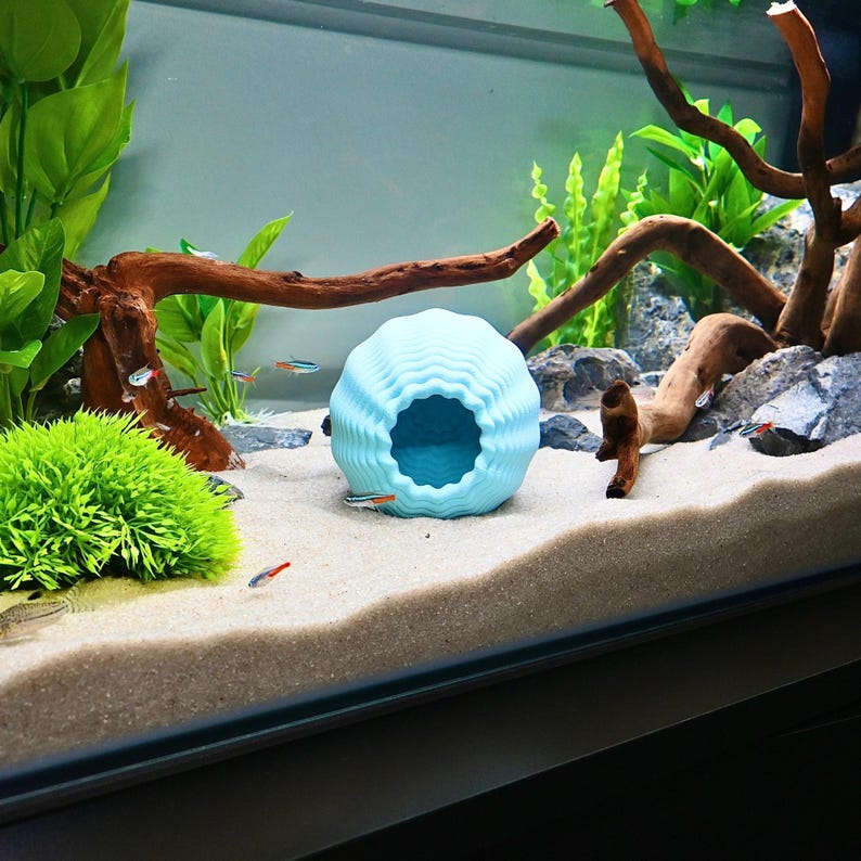 May include: A pale blue spherical aquarium ornament with a textured, wavy surface and a large opening. The ornament is in an aquarium with sand, plants, and driftwood. Small fish swim around the ornament.