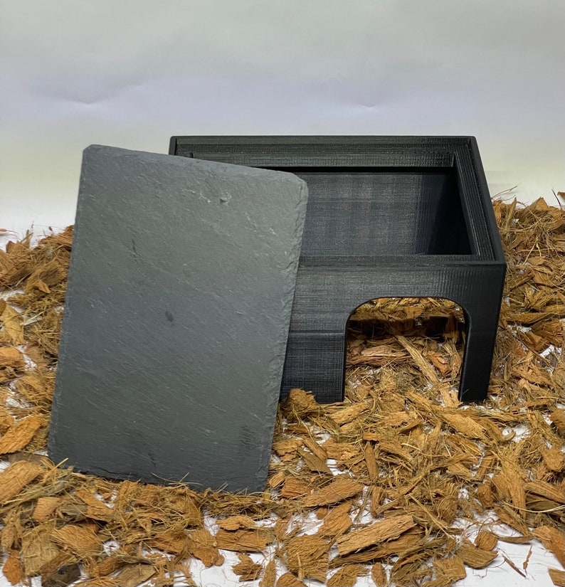 May include: A black 3D printed hideaway for small pets, with a removable slate roof. The hideaway is sitting on a bed of brown coconut fiber.