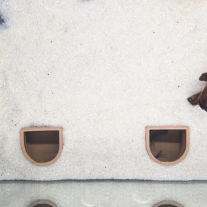 May include: Two terracotta-colored, semi-circular aquarium caves are embedded in white sand. The caves have a rounded top and a flat bottom, creating a small, sheltered space. The sand has a fine, granular texture, and a piece of driftwood is visible.