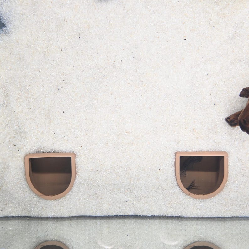 May include: Two terracotta-colored, semi-circular aquarium caves are embedded in white sand. The caves have a rounded top and a flat bottom, creating a small, sheltered space. The sand has a fine, granular texture, and a piece of driftwood is visible.