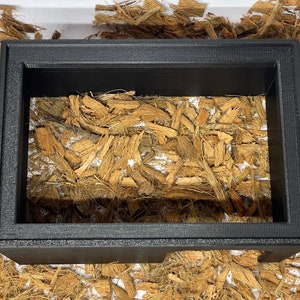 May include: A black picture frame with a wood-like material inside. The frame is sitting on a white surface.