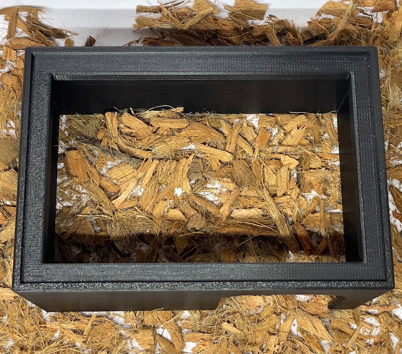 May include: A black picture frame with a wood-like material inside. The frame is sitting on a white surface.