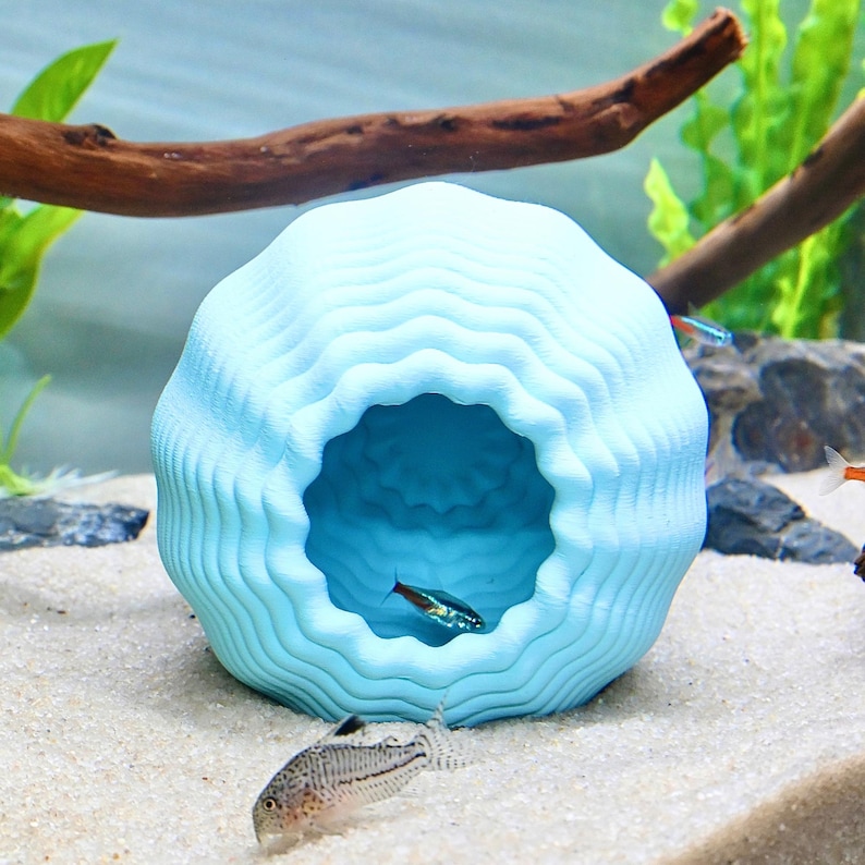 May include: A light blue, wave-patterned aquarium cave. The cave has a large opening and is surrounded by sand, plants, and fish. The cave is designed to provide shelter for fish and other aquatic creatures.