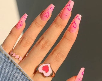 Könnte beinhalten: Lange, rosa Press-on-Nägel mit einem rosa Herz-Design. Die Nägel werden auf einer Hand mit einem goldenen Ring mit einem rosa Herz-Design gezeigt.