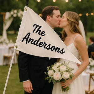 May include: A white flag with the words "The Andersons" in black script is held in front of a kissing couple. The bride holds a bouquet of white roses. The scene is outdoors with string lights.