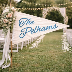 Pode incluir: Uma bandeira branca com as palavras "The Pelhams" em azul é exibida em um casamento ao ar livre. A bandeira é decorada com flores e fitas. Cadeiras brancas alinham o corredor, que é coberto por pétalas de flores.