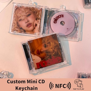 May include: A clear plastic keychain with a mini CD inside. The CD has a pink background with the word "a!e" in black text. The keychain is a custom design and features a photo of a woman on the front and back. The keychain also has a small NFC chip that can be used to play music.