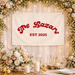 Peut inclure: Une bannière crème avec le texte "The Lazars EST 2025" en script rouge. La bannière est suspendue devant un rideau blanc, avec des compositions florales et des bougies sur une table.