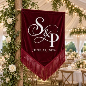 May include: A deep red velvet pennant banner featuring white stylized lettering of the initials S & P and the date JUNE 29, 2026. The triangular banner is suspended from a wooden rod and finished with a long matching red fringe along the bottom edge.