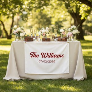 Könnte beinhalten: Ein Tisch für eine Hochzeitsfeier im Freien, bedeckt mit einer cremefarbenen Tischdecke. Der Tisch ist mit Blumenarrangements, Sektgläsern und kleinen Holzkisten geschmückt. Ein weißes Banner zeigt den Text "The Williams" und das Datum "07/02/2026".