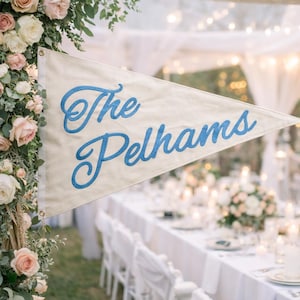 Puede incluir: Una bandera de banderín color crema con las palabras "The Pelhams" bordadas en escritura azul. La bandera se muestra en un evento al aire libre, con una mesa larga preparada para una comida en el fondo. También se ven arreglos florales y luces de cadena.