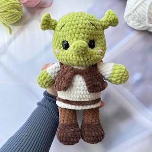 May include: A handmade green crocheted Shrek doll with black eyes, a brown vest, and brown boots. The doll has a white shirt and green arms and head. The doll is made of soft yarn and is a cute, whimsical toy.