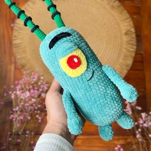 May include: A plush toy shaped like a small, cylindrical creature with a teal body, two long green antennae with black rings, a single large yellow eye with a red pupil, and a black embroidered eyebrow and mouth, crafted from soft yarn.