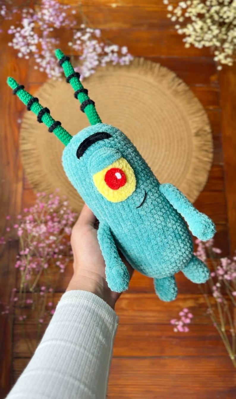 May include: A plush toy shaped like a small, cylindrical creature with a teal body, two long green antennae with black rings, a single large yellow eye with a red pupil, and a black embroidered eyebrow and mouth, crafted from soft yarn.