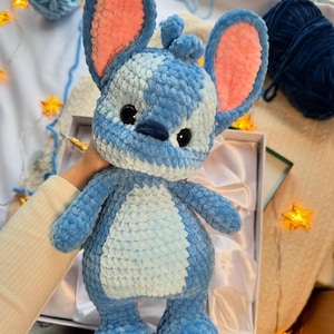 May include: A handmade blue plush toy with large ears and a white belly. The toy has black eyes and a small nose. The ears are lined with pink. The toy is made of a soft, textured material and is presented in a gift box.