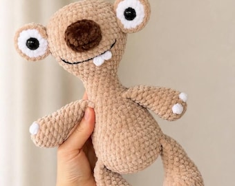 May include: A handmade, tan-colored crochet creature with large, round eyes, a brown nose, and two small white teeth. The plush toy has small arms and legs with white accents on the feet and hands. The toy is held up by a hand.